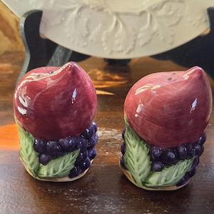 May include: A pair of ceramic salt and pepper shakers. Each shaker is shaped like a red fruit with a cluster of purple grapes at the base. Green leaves are visible. The shakers have small holes on top for dispensing.