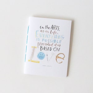 May include: A white card with blue and light blue text that reads "in the arts, as in life, everything is possible provided it is based on love". The card has a blue, white, and yellow striped border and a small illustration of a button, scissors, and a ring.