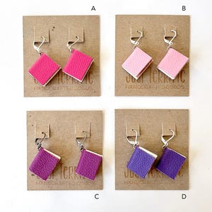 May include: Four pairs of leather book earrings on display cards. The earrings are in pink, light pink, purple, and lavender. Each card has the text "Just Terrific Handcrafted Goods".