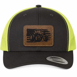 May include: A black and neon yellow trucker hat with a rectangular brown leather patch on the front. The patch features a laser-engraved image of a tractor superimposed over an American flag. The hat has a black brim and a snapback sticker.