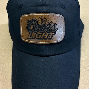 May include: A black baseball cap featuring a rectangular brown leather patch centered on the front. The patch displays the Coors Light logo in black, including the mountain graphic and the text Coors Light, with visible stitching around the edges.