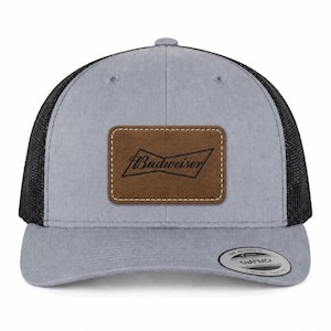 May include: A grey baseball cap with black mesh back panels and a brown rectangular leather patch on the front. The patch features the black Budweiser logo. A round sticker on the brim reads The Authentic Snapback.