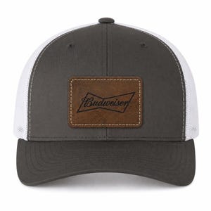 May include: A charcoal gray baseball cap with white mesh side and back panels. The front features a rectangular brown faux leather patch with the black Budweiser logo and text stitched in the center. The cap has a curved brim and a button top.