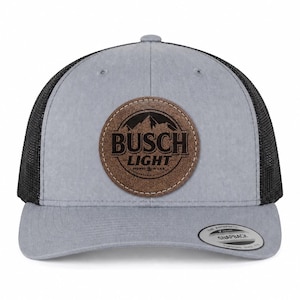 May include: A grey baseball cap with black mesh back panels and a snapback closure. The front features a circular brown leather patch with the black text BUSCH LIGHT and a mountain graphic. A round sticker is on the brim reading THE AUTHENTIC SNAPBACK.
