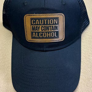 May include: A black baseball cap with a mesh back and a rectangular brown patch on the front. The patch features the black text CAUTION MAY CONTAIN ALCOHOL in a bold, sans-serif font, enclosed within a thin black border on the leather-like material.