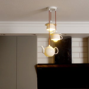 May include: A quirky pendant light fitting with three off-white ceramic shades: a teapot and two teacups. The shades hang from brown cords and a white ceiling mount. The kitchen setting includes grey cabinets and white tiles.