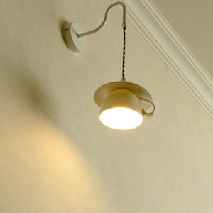 Op de afbeelding: Een off-white wandlamp met een lampenkap in de vorm van een theekopje. Het licht hangt aan een gebogen arm die aan een cirkelvormige wandbevestiging is bevestigd. Het snoer is zwart en wit. Het licht is aan.