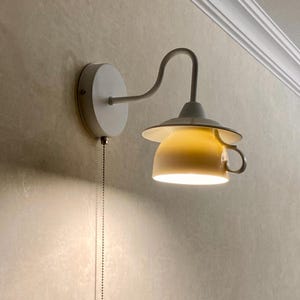 Może przedstawiać: Biała lampa ścienna z zakrzywionym ramieniem i żółtym kloszem w kształcie filiżanki do herbaty. Lampa ma łańcuszek i jest zamontowana na teksturowanej ścianie. Światło świeci w dół.
