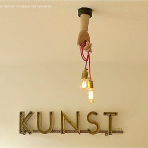 May include: A unique light fixture featuring a wooden hand holding two Edison-style light bulbs with red and purple cords. Below, the word "KUNST" is spelled out in metal letters on the wall. The fixture is mounted on a white ceiling.
