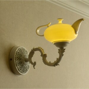 Può includere: Una lampada da parete decorativa a forma di teiera. La lampada presenta un paralume in vetro giallo luminoso, un manico curvo e una base decorativa bianco sporco. La lampada è montata su una parete beige. Misure in centimetri.