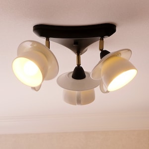 Può includere: Un moderno lampadario a soffitto con tre paralumi bianchi a forma di tazza. L'apparecchio ha una base nera e dettagli color ottone. Le luci sono accese, emettendo un bagliore caldo.