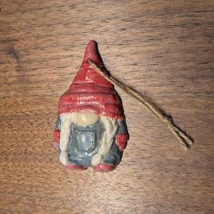 May include: A red and white gnome ornament with a gray outfit and white beard. The gnome is wearing a red hat and has a string attached to the top of the hat.