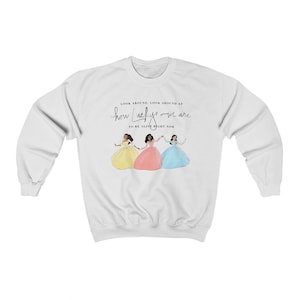Peut inclure: Sweat-shirt blanc avec une illustration de trois femmes en robes colorées, se tenant par la main. Le texte "LOOK AROUND, LOOK AROUND AT how LUCKY we are TO BE ALIVE RIGHT NOW" est imprimé au-dessus de l'illustration en script noir.