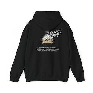 Puede incluir: Sudadera con capucha negra con un diseño de frasco de perfume dorado y el texto "IANNEASHE". Las palabras "Sorry Baby" están escritas en blanco encima del frasco. Debajo, nombres de ciudades: Viena, Londres, París, Barcelona, Ámsterdam, Roma, Berlín, Moscú, Ginebra.