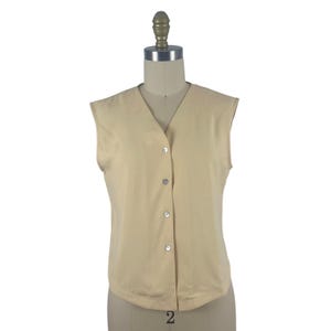 May include: A sleeveless beige vest featuring a V-neckline and a front button closure with four small, round, light-colored buttons. The garment is constructed from a solid-colored fabric and has a straight hemline, displayed on a mannequin.