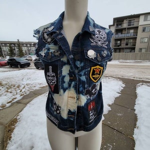May include: A blue denim vest with various patches and pins. The patches include a skull, a sword, a star, and a cloud. The pins are mostly silver and black.
