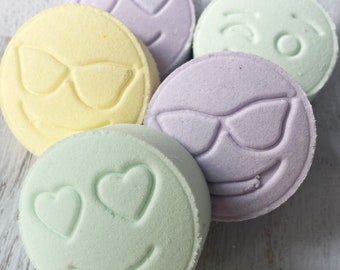May include: A collection of colorful, round bath bombs with embossed smiley faces. The bath bombs are in shades of yellow, lavender, and green, each with a different expression, including sunglasses, hearts for eyes, and a winking face.