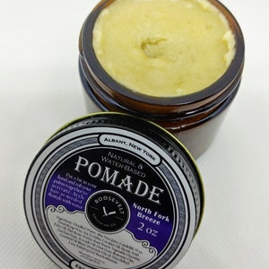 May include: A 2 ounce jar of natural water-based pomade labeled "Roosevelt Grooming Co. North Fork Breeze" with a black and white label on a brown jar. The jar is open and the pomade is a light yellow color.