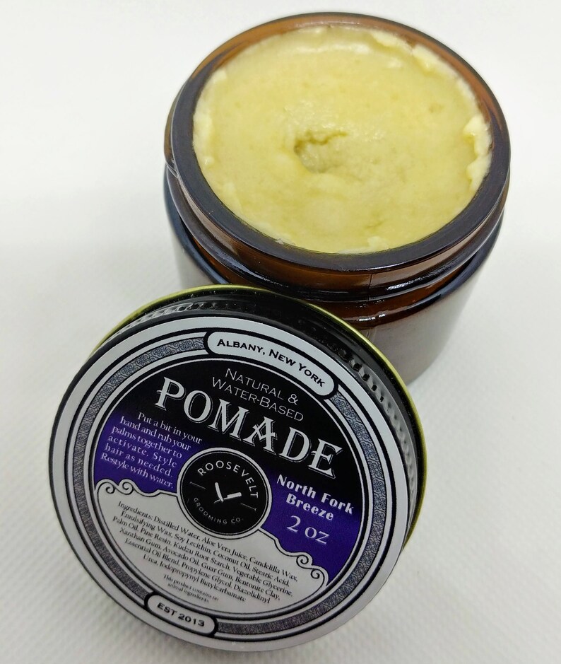May include: A 2 ounce jar of natural water-based pomade labeled "Roosevelt Grooming Co. North Fork Breeze" with a black and white label on a brown jar. The jar is open and the pomade is a light yellow color.