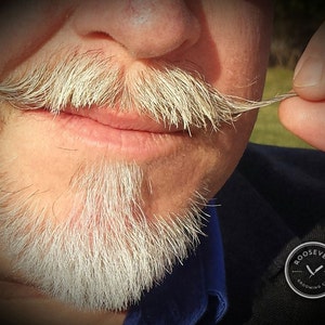 Puede incluir: Un hombre con barba y bigote blancos, sujetando el extremo de su bigote con la mano derecha. El hombre lleva una camisa azul y una chaqueta negra con un logotipo que dice "Roosevelt Grooming Co."