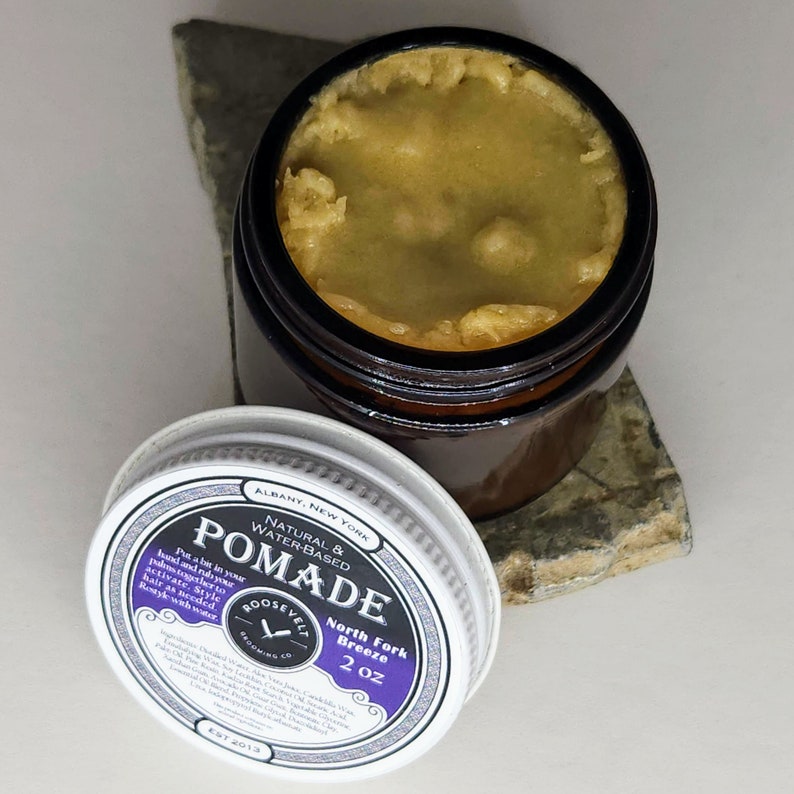 May include: A 2 ounce jar of natural water-based pomade labeled "North Fork Breeze" by Roosevelt Grooming Co. The jar is brown glass with a white lid. The pomade is a light yellow color.