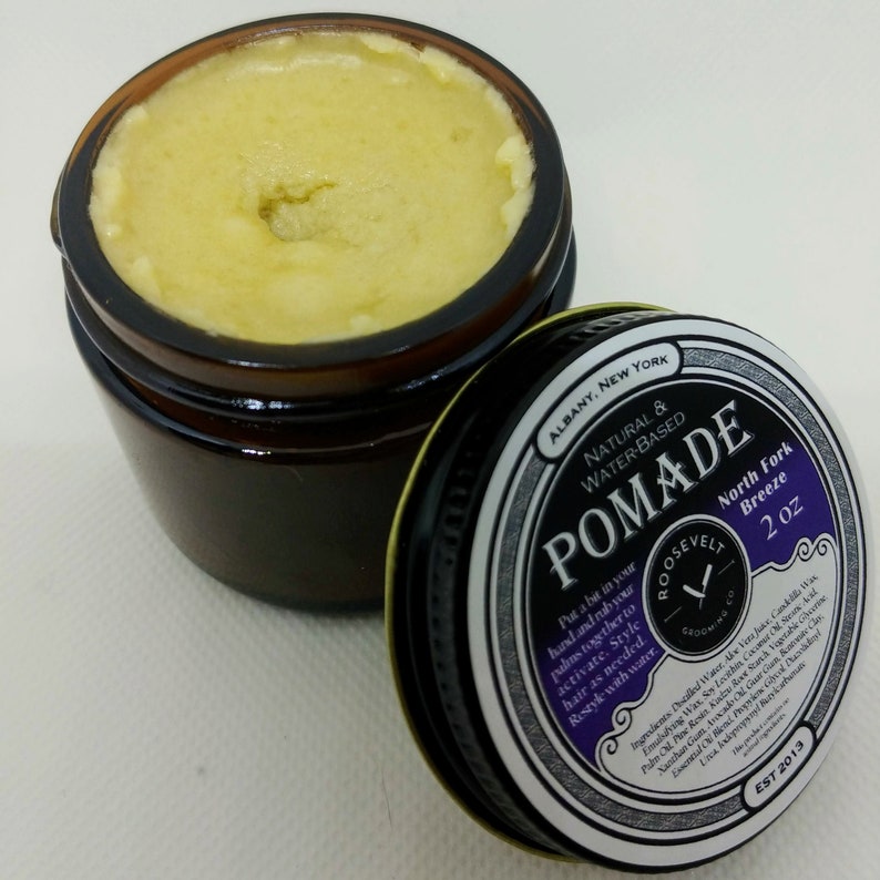 May include: A 2 ounce jar of natural water-based pomade labeled "POMADE" with the scent "North Fork Breeze". The jar is brown glass with a black lid. The label is black with white and gold lettering and a purple background. The label also includes a logo with a razor blade and the text "ROOSEVELT GROOMING CO. EST 2013."