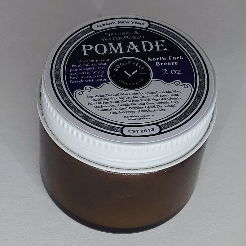 May include: A white jar with a black and silver label that reads "Roosevelt Grooming Co. Pommade North Fork Breeze 2 oz. Est 2013". The label also lists the ingredients for the product.