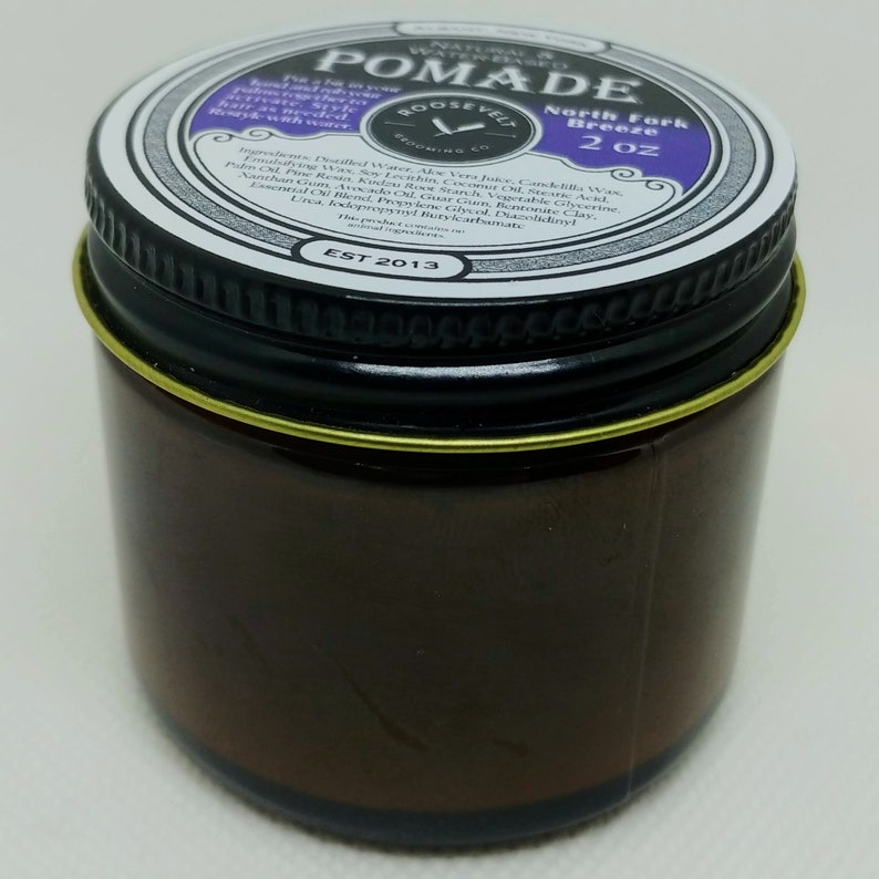 May include: A brown glass jar with a black lid and a gold band around the top. The lid has a white label with the text "NATURAL & WATER-BASED POMADE" in black lettering. The label also includes the text "ROOSEVELT GROOMING CO. EST 2013" and "North Fork Breeze 2 oz".