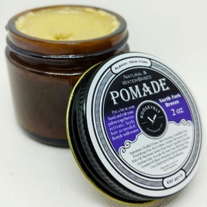 May include: A 2 ounce jar of natural water-based pomade labeled "Roosevelt Grooming Co. Pommade North Fork Breeze". The jar is brown with a black lid and a purple and white label. The label has a vintage design with a razor blade logo and the text "EST 2013".