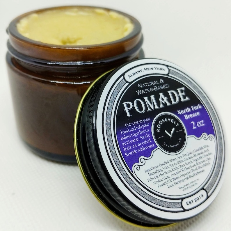 May include: A 2 ounce jar of natural water-based pomade labeled "Roosevelt Grooming Co. Pommade North Fork Breeze". The jar is brown with a black lid and a purple and white label. The label has a vintage design with a razor blade logo and the text "EST 2013".