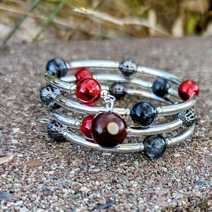 May include: A silver bracelet with multiple strands of beads. The beads are red, black, and brown. The bracelet is on a brown, rocky surface.