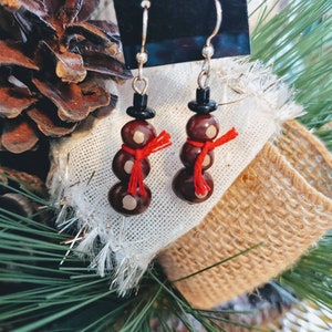 May include: Snowman earrings with silver hooks. Each earring features three brown beads with white dots, a black bead hat, and a red scarf. The earrings are displayed on a white cloth with a burlap ribbon and pine needles in the background.
