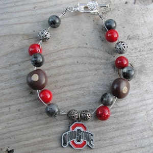 Könnte beinhalten: Ein rotes und graues Perlenarmband mit einem silbernen Anhänger mit dem Ohio State Logo. Der Anhänger ist mit einer silbernen Schlaufe am Armband befestigt.