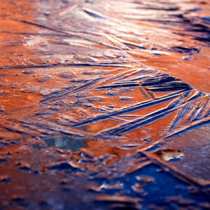 Op de afbeelding: Abstracte macrofotografie van een bevroren oppervlak. De afbeelding toont een mix van oranje en blauwe tinten, met ingewikkelde patronen van ijskristallen. De textuur is ruw en ongelijkmatig, wat een visueel interessante compositie creëert.