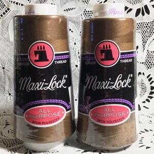 以下が含まれることがあります： オーバーロックミシン用のブラウンのMaxi-Lockオールパーパススパンポリエステル糸の糸巻き2個。糸巻きには、白文字の黒ラベルと、ミシンをモチーフにした赤い円形が描かれています。