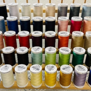 Könnte beinhalten: Eine Sammlung von vierzig Rollen Gutermann-Nähgarn, die auf einem Holzständer angeordnet sind. Die Garnrollen weisen eine Vielzahl von Volltonfarben auf, darunter Schwarz, Weiß, Blau, Rot, Grün, Gelb, Rosa, Braun und Grau, jeweils mit Etikett.