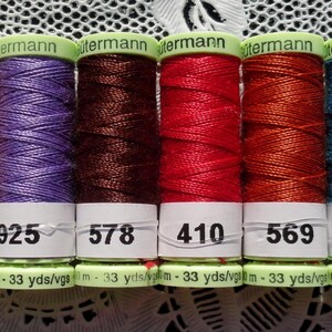 May include: A collection of six spools of Gütermann thread in various colors: gold, purple, brown, red, orange, and teal. Each spool has a white label with a number and the text "33 yds". The spools are arranged in a row.