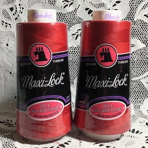 May include: Two spools of red Maxi-Lock thread. Each spool has a black label with white text, including "Maxi-Lock", "Serger Thread", and "All Purpose Spun Polyester". The labels also feature a pink circle with a sewing machine icon.