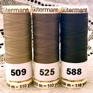 Könnte beinhalten: Drei Spulen Gütermann-Garn in Beige, Grau und Schwarz. Jede Spule hat ein weißes Etikett mit dem Gütermann-Logo und der Nummer 509, 525 bzw. 588. Die Spulen sind mit 100 m - 110 yds gekennzeichnet.
