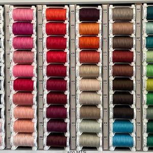 Pode incluir: Uma exibição de bobinas de linha coloridas dispostas em uma grade. As bobinas são em várias tonalidades de preto, cinza, azul, rosa, vermelho, laranja, marrom, verde e amarelo. O texto "600 MTS" é visível na parte inferior da imagem.