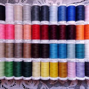 May include: A collection of colorful spools of thread, arranged in neat rows. The threads are in a variety of colors, including black, white, pink, blue, and orange. Each spool has a label with text. The spools are on a white lace surface.