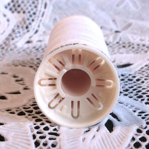 May include: A white plastic spool with a hole in the center and a series of small holes around the edge. The spool is designed to hold thread.