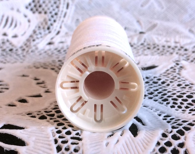 May include: A white plastic spool with a hole in the center and a series of small holes around the edge. The spool is designed to hold thread.