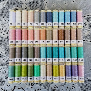 Puede incluir: Una colección de coloridas bobinas de hilo Gutermann, dispuestas en filas ordenadas. Los hilos vienen en una amplia gama de colores, desde pasteles hasta tonos vibrantes. Cada bobina está etiquetada con un número. Los hilos son de 100 metros.