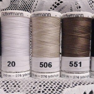 May include: Five spools of Gutermann thread in various colors: black, white, beige, brown, and dark brown. Each spool has a white label with a number and the Gutermann logo. The thread is wound on cylindrical spools, with the numbers 10, 20, 506, 551, and 594 visible.