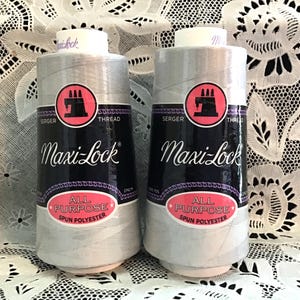 以下が含まれることがあります： Maxi-Lockのオーバーロック糸の2つのスプール。スプールは、黒とピンクのラベルが付いたライトグレーです。ラベルには「Maxi-Lock」と「All Purpose Spun Polyester」と記載されています。糸は白いスプールに巻かれています。