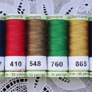 May include: Six spools of Gütermann thread in various colors: dark blue, red, brown, green, gold, and purple. Each spool has a white label with a number and the text "30 m - 33 yds". The thread is displayed on a white lace surface.