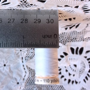 May include: A white spool of thread with a label that reads "110 yds/10 m - 110 yds/10 m". The spool is measured against a ruler.