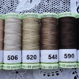 Puede incluir: Seis carretes de hilo Gütermann en varios tonos de marrón, desde beige claro hasta marrón oscuro. Cada carrete tiene una etiqueta blanca con un número y el texto "33 yds/vgs". Los carretes están dispuestos en fila.
