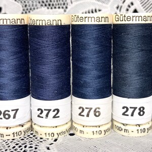 Könnte beinhalten: Sechs Spulen dunkelblauen Fadens mit weißen Etiketten. Die Etiketten tragen den Text "Gutermann" und die Nummern "238", "267", "272", "276", "278" und "279". Jede Spule hat die Maße "100 m - 110 yds".